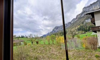 A view through a window onto a green landscape with mountains in the background. The sky is cloudy and there are some trees in the vicinity.