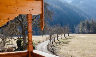 A beautiful balcony with wooden embellishments and a view of a picturesque mountain landscape. In the background, snow-capped mountains and a peaceful path can be seen.