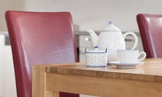 A teapot and cups made of ceramic with blue dots are on a wooden table. In the background, red chairs can be seen.