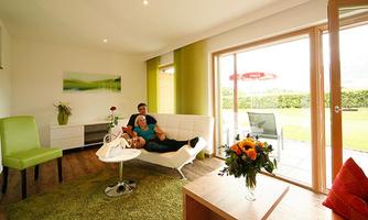 A cozy living space with modern furniture and a large window front. A couple relaxes on a white sofa, surrounded by fresh flowers and a green carpet.