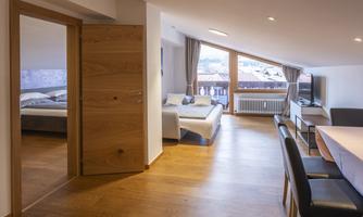 A modern, bright apartment with wooden flooring and large windows. The space is furnished with a cozy sofa, a dining area, and access to a bedroom.