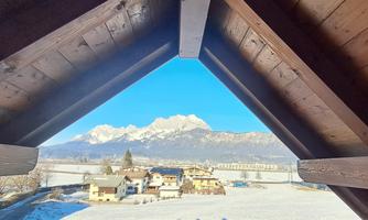 A view from a wooden frame of a snowy valley with houses and snow-covered mountains in the background. The sky is bright blue and clear.