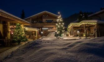 A winter landscape with snow-covered ground and festively decorated buildings. In the foreground stands a illuminated Christmas tree.