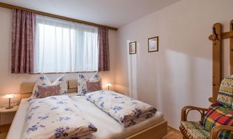A cozy bedroom with a double bed and colorful cushions. Soft curtains and a wicker chair give the room an inviting atmosphere.