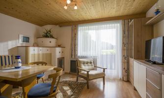 A cozy living space with a wooden ceiling and large windows. The room is furnished with a dining table, chairs, and a television.