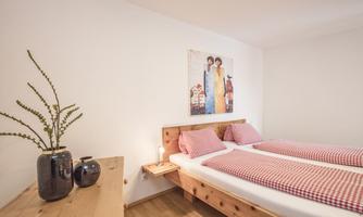 A cozy bedroom with two beds and checkered bedspreads. A colorful painting hangs on the wall, and a plant along with vases adorn the dresser.