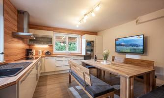 A modern kitchen with a large dining table and a bright, friendly atmosphere. The room features a combination of wooden and white furniture, as well as a window with a view of greenery.
