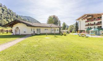 A well-kept garden with lawns and a terrace. In the background, several buildings and wooded mountains can be seen.