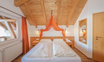 A cozy bedroom with a large double bed and a wooden ceiling. The walls are painted light, and there are orange curtains as well as a mirror.
