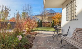 An inviting garden with colorful plants and a cozy bench. In the background, light mountains and a blue sky can be seen.