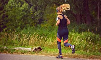 A woman jogs along a path, surrounded by green nature. She is wearing athletic clothing and headphones.