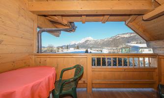 A cozy balcony with a table and a chair. In the background, snow-covered mountains and a clear blue sky can be seen.