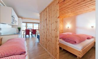 A cozy bedroom with wooden walls and two beds. In the background, there is a bright living area with a dining table and chairs.