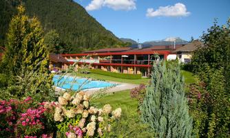 A beautiful hotel with a pool, surrounded by colorful flowers and green trees. In the background, mountains and a clear sky can be seen.