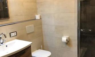 A modern bathroom with a shower, a sink, and a toilet. The walls are light tiled and the floor is dark.