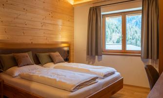 A cozy bedroom with wood paneling and a large window. The room features a double bed and a desk.