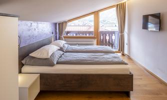 A modern bedroom with two beds and large windows. The room features hardwood floors and simple, elegant furnishings.