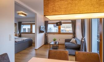 A modern living and dining room with wooden flooring. The room is bright and warmly furnished, with a sleeping area, a sofa, and a dining table.