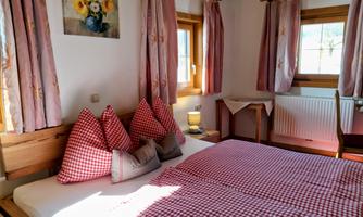 A cozy bedroom with a double bed and checkered pillows. The walls are decorated with pink curtains and a blooming painting.