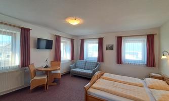 A cozy bedroom with two beds, a table, and a sofa. There are windows with curtains that provide plenty of daylight.