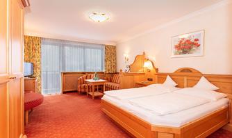 A cozy hotel room with a double bed and a living area. The walls are in warm wood tones and large windows provide plenty of light.
