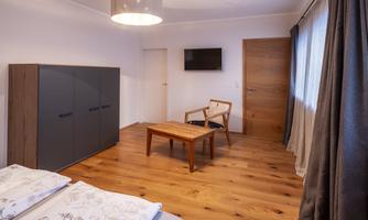 A modern bedroom with wooden flooring and a large window. There is a bed, a table, and a television.