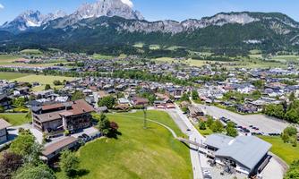 A picturesque landscape with a small village in the foreground and majestic mountains in the background. The surroundings are green and inviting under a clear sky.