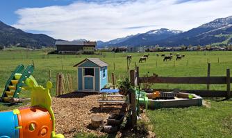 A playground with colorful play equipment and a small house. In the background, green meadows and cows can be seen in front of a mountain range.