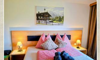 A cozy bedroom with two beds, decorated with red pillows and a beautiful landscape photograph on the wall. There are also nightstands with lamps and curtains that create a homely atmosphere.