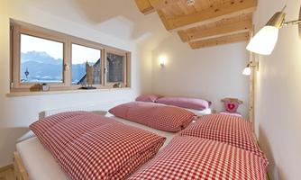 A cozy bedroom with a wooden beam ceiling and large windows. The beds are equipped with checkered pillows and blankets.
