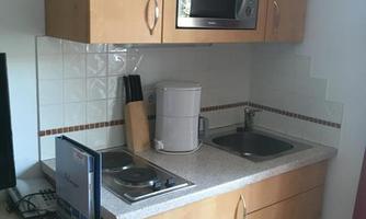 A small kitchen with wooden furniture, microwave, and two sinks. On the left, there is a television and some kitchen appliances are visible.