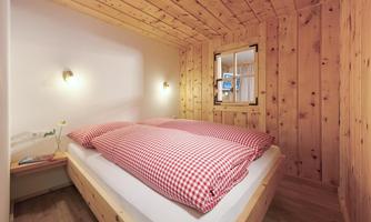 A cozy bedroom with wooden paneling and a double bed. The bedding is red and white checkered, and there is a window with natural light.