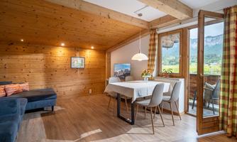 A cozy living room with wooden walls and a dining area. Large windows offer a view of the surrounding landscape.