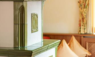 A traditional tiled stove with green and white tiles stands in a cozy room. In the background, cushions and curtains can be seen, creating an inviting atmosphere.