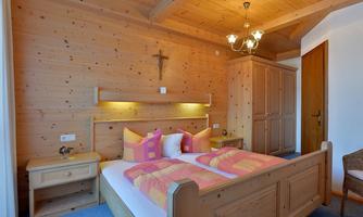 A cozy bedroom with wooden paneling, a double bed, and colorful cushions. The room is bright and inviting, with a small nightstand and a wardrobe.