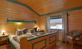A cozy bedroom with wood paneling and colorful accents. There is a large bed, an armchair, and a view of the outside from the window.