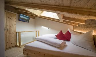 A cozy and rustic-looking bedroom with wooden ceilings and a double bed. A television and large windows create a bright atmosphere.