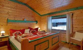 A cozy wooden bedroom with a double bed and colorful pillows. Next to the bed is a rattan chair, and the window offers a view outside.