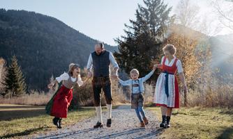 A family in traditional clothing happily walks along a path. In the background, trees and mountains can be seen, creating a picturesque landscape.