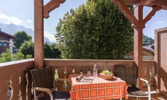 A cozy balcony with a table and two chairs. The table is set with a checkered tablecloth and offers a view of trees and the mountains with the Zahmer Kaiser in the background.