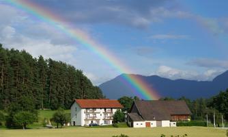 A beautiful rainbow over two traditional houses in a rural setting. In the background, green forests and mountains can be seen.