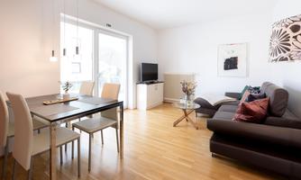 A modern living room with a comfortable sofa and a glass table. There is a dining area with a table and chairs, as well as large windows that let in lots of light.