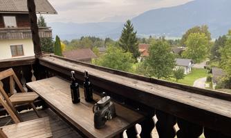 A balcony with wooden chairs and a table. On the table, there are two bottles and an old camera, while the landscape with trees and mountains in the background is visible.