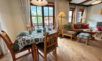 An inviting living room with a table and chairs, surrounded by large windows. A cozy seating area with cushions and bright curtains creates a friendly atmosphere.