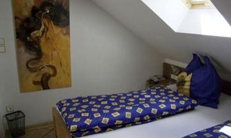 A simple bedroom with two beds, blue bed covers, and a window in the roof area. An abstract artwork is hanging on the wall.