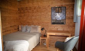 A cozy bedroom with wooden paneled walls. There is a bed, a table, a chair, and a piece of art on the wall.