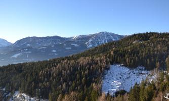A picturesque mountain landscape with snowy peaks and wooded slopes. The sky is clear and blue.