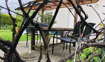 A garden with a table and chairs, surrounded by plants. In the background, a house with a covered terrace can be seen.