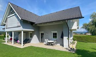 A modern house with a beautiful garden. There is a terrace with furniture and a clear blue sky.