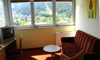 A cozy living space with a sofa, a small table, and a television. Large windows provide a view of the green landscape outside.